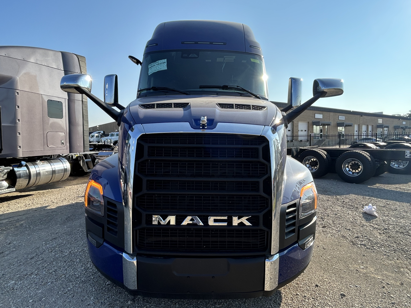Truck front view showing Mack grille, large chrome bars, and blue cab with side mirrors; headlights and badge visible.
