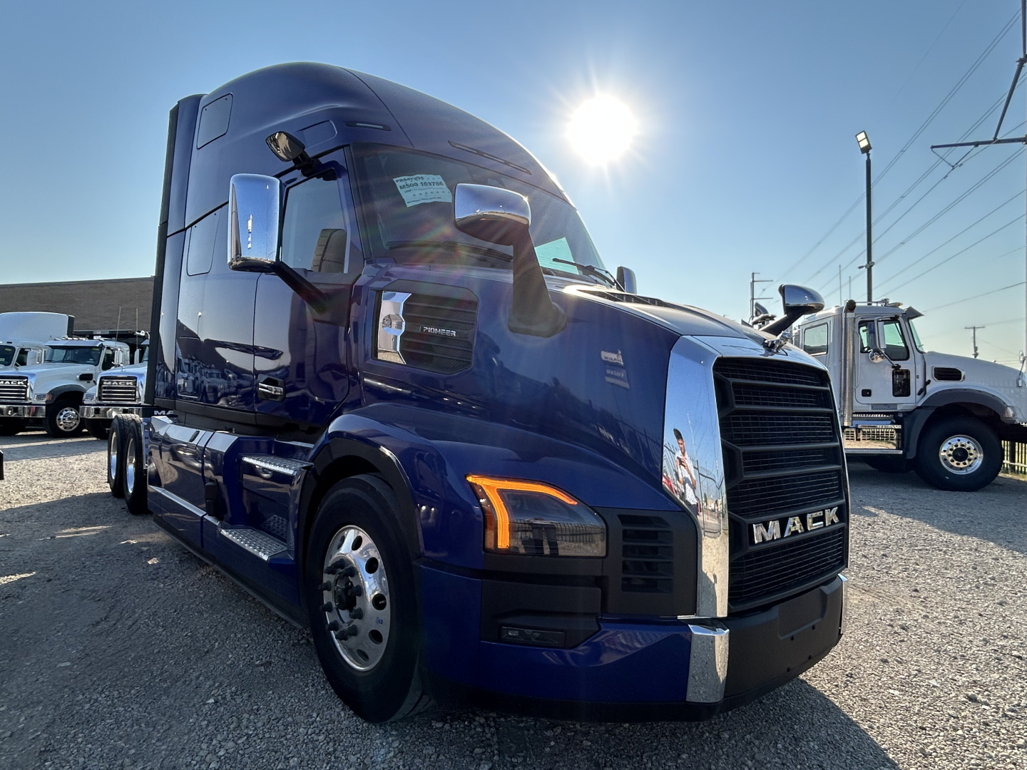 Blue Mack PR64T76H truck cab front view with bold grille and headlights, side mirrors, and sun glare.