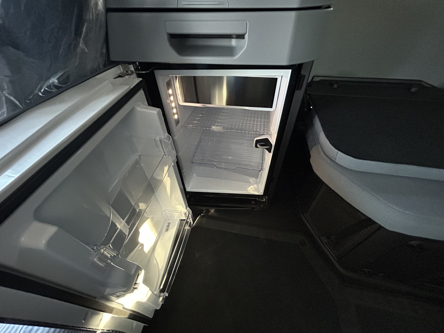 Interior view of a new Mack PR64T76H truck cab showing an open small refrigerator/freezer unit with light inside and a passenger seat nearby.