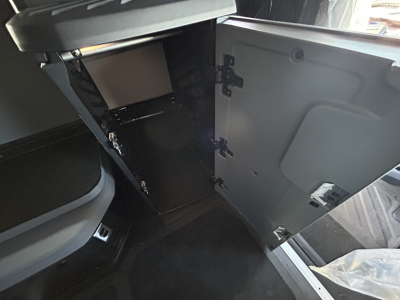 Interior cab storage area of a new 2027 Mack PR64T76H truck showing open cabinet with shelves and a door, dark gray interior, and a portion of the sleeper area.