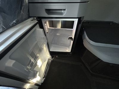 Interior view of a new Mack PR64T76H truck cab showing an open small refrigerator/freezer unit with light inside and a passenger seat nearby.