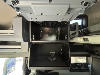 Cab interior of a 2027 Mack PR64T76H showing open storage compartments under the dashboard with black plastic drawers and metal mounting brackets.