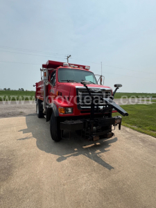 Red Sterling L7500 dump truck front view with plow attachment and light bar. Windshield shows faint watermark. Landscape background with road and grass.