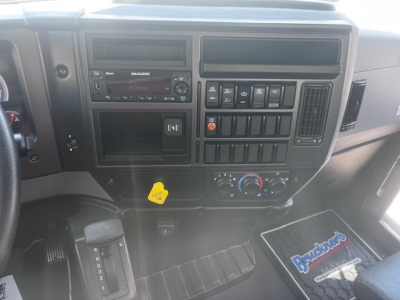 Dashboard center console of a Mack MD6 truck showing stereo radio, various switches, climate controls, and a yellow tag; vinyl floor mat and storage compartments visible.