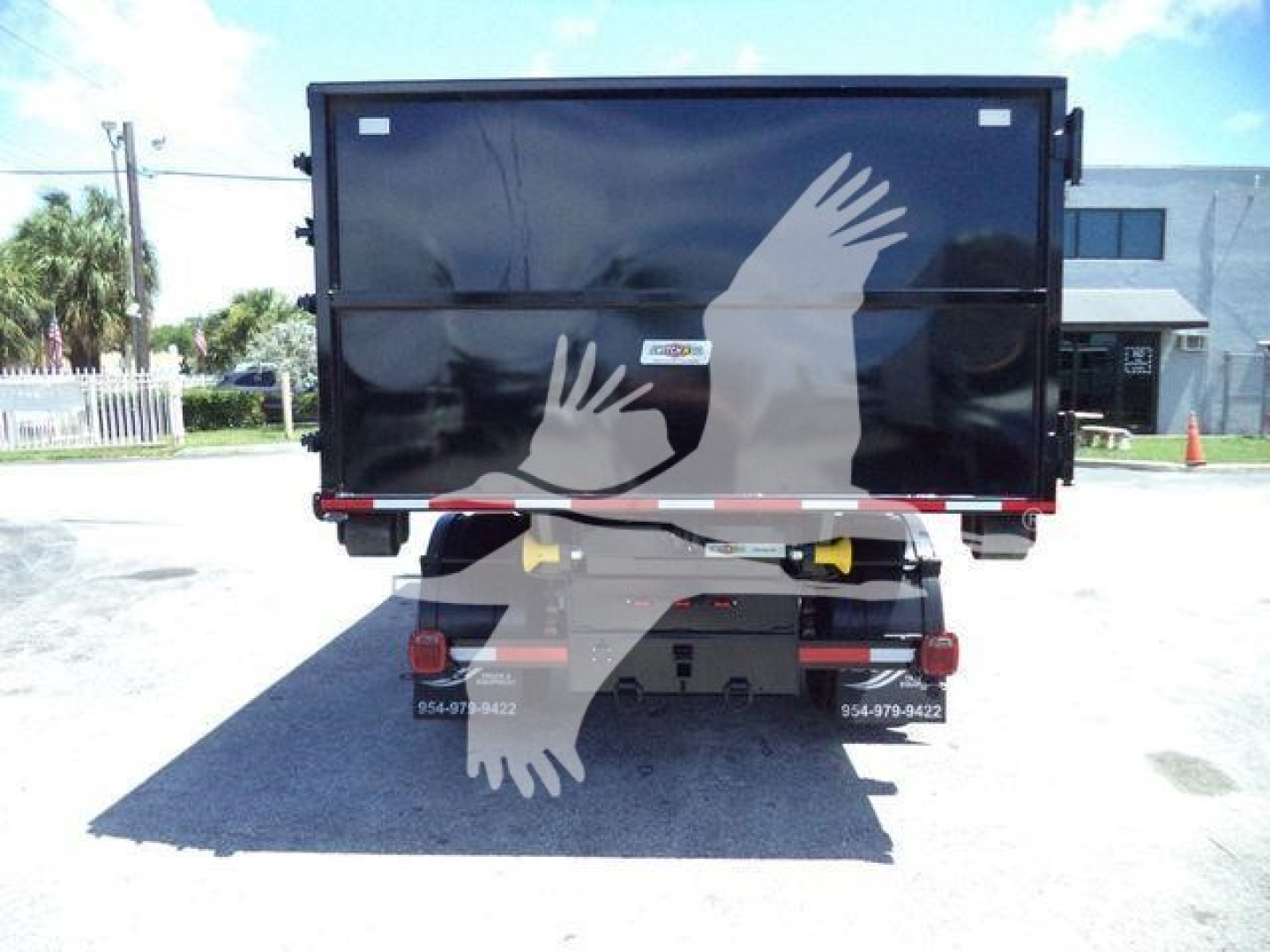 Back view of a black roll-off truck body with a yellow hydraulic component, bumper, and rear lights. faint white silhouettes of hands on the tailgate. No legible serials or labels visible.