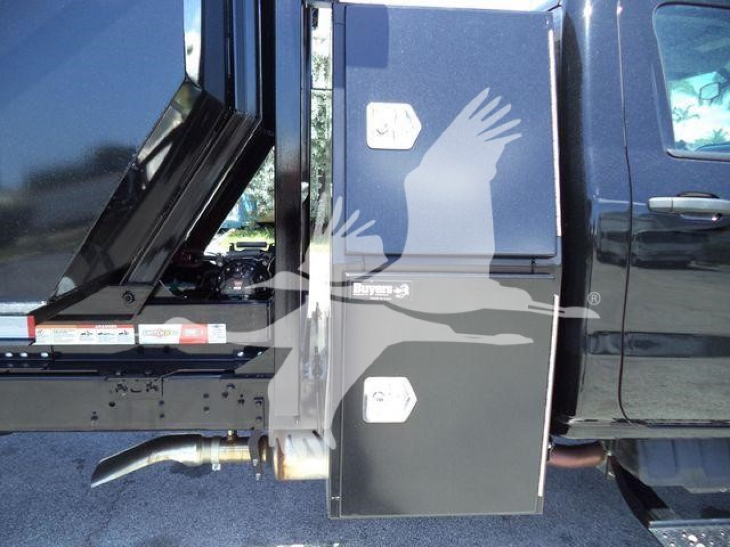Truck side view showing a black metal toolbox on the cab with a raised dump bed behind; faint text labels on small panels.