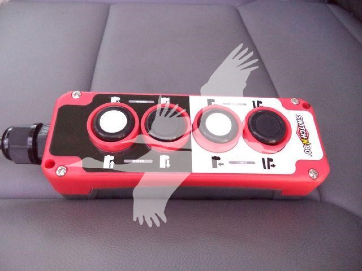 Control panel with red housing showing four circular ports and labels. Left side has a black connector. The panel has white and black labeling, including small symbols near each port.
