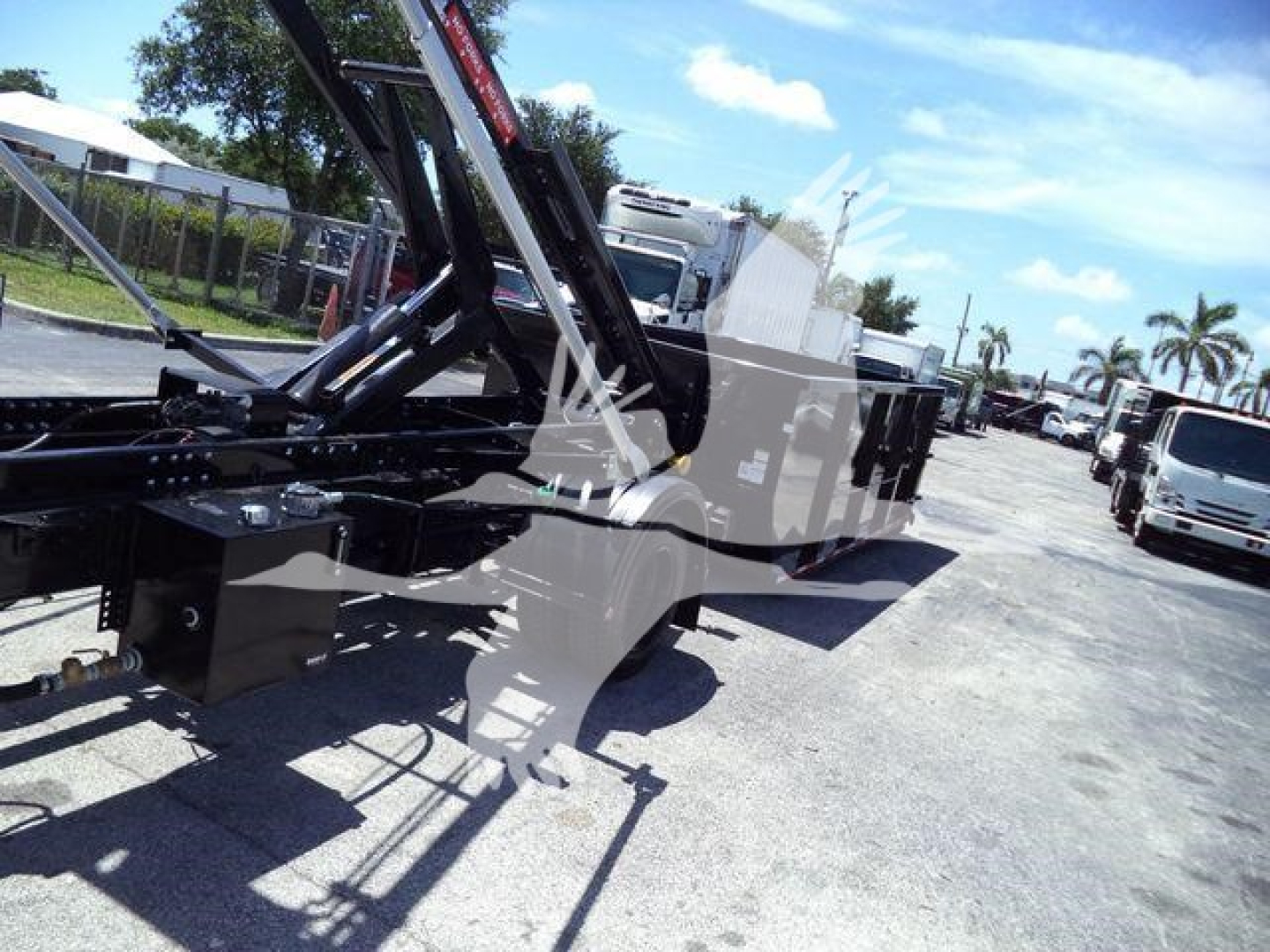Equipment trailer showing a large roll-off truck body with hydraulic arms and black metal frame, parked outdoors with other trucks in background.