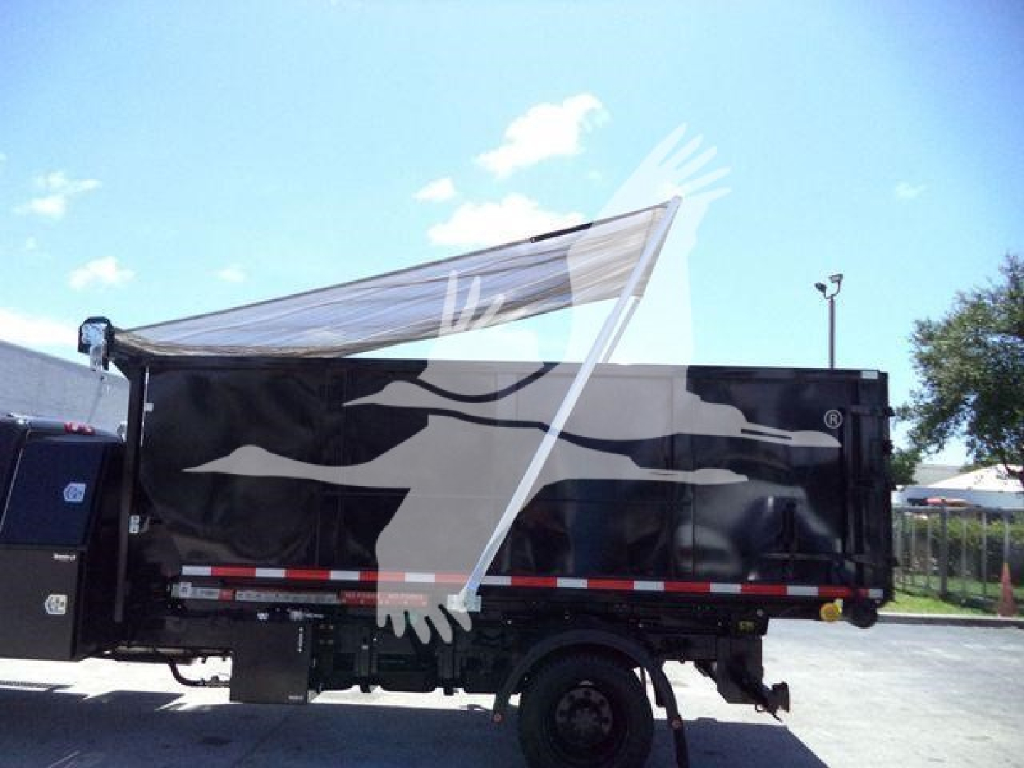 Dump truck side view showing a black dump body with white graphics and a partially extended tarp on a sunny day.