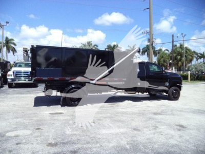 Side view of a black dump truck with a large cargo bed on a dual rear wheel chassis. Stickered deck and side rails visible under bright sky.