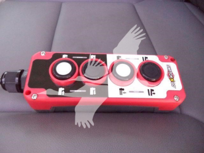 Control panel with red housing showing four circular ports and labels. Left side has a black connector. The panel has white and black labeling, including small symbols near each port.