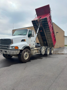 Truck cab with dumping bed raised on a white Sterling LT9500 dump truck, logo visible, multiple axles, front grille, and red dumped bed.