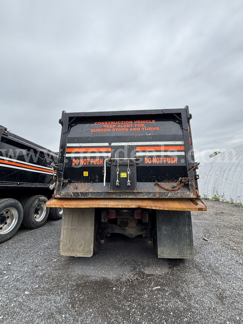 Back view of a Mack Granite dump truck bed with warning decals and hydraulic tailgate, rusty low area, wheels and chassis visible.