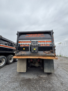 Back view of a Mack Granite dump truck bed with warning decals and hydraulic tailgate, rusty low area, wheels and chassis visible.