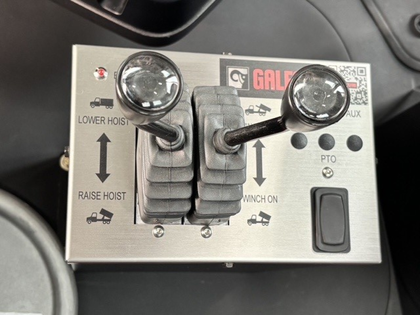 Control panel with two joysticks and labeled buttons: LOWER HOIST, RAISE HOIST, CUT? arrows, WINCH ON, PTO. Brand label GAL? with a QR code. Metal faceplate with two levers and surrounding indicators.