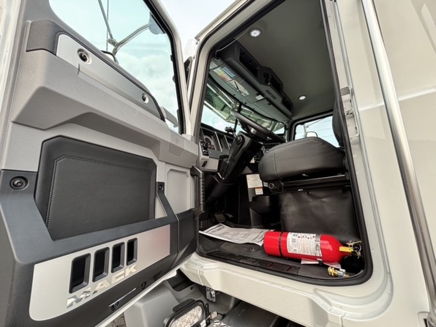 Truck cab interior showing open door and steering wheel of Mack GR64B roll-off truck with a fire extinguisher on the floor.