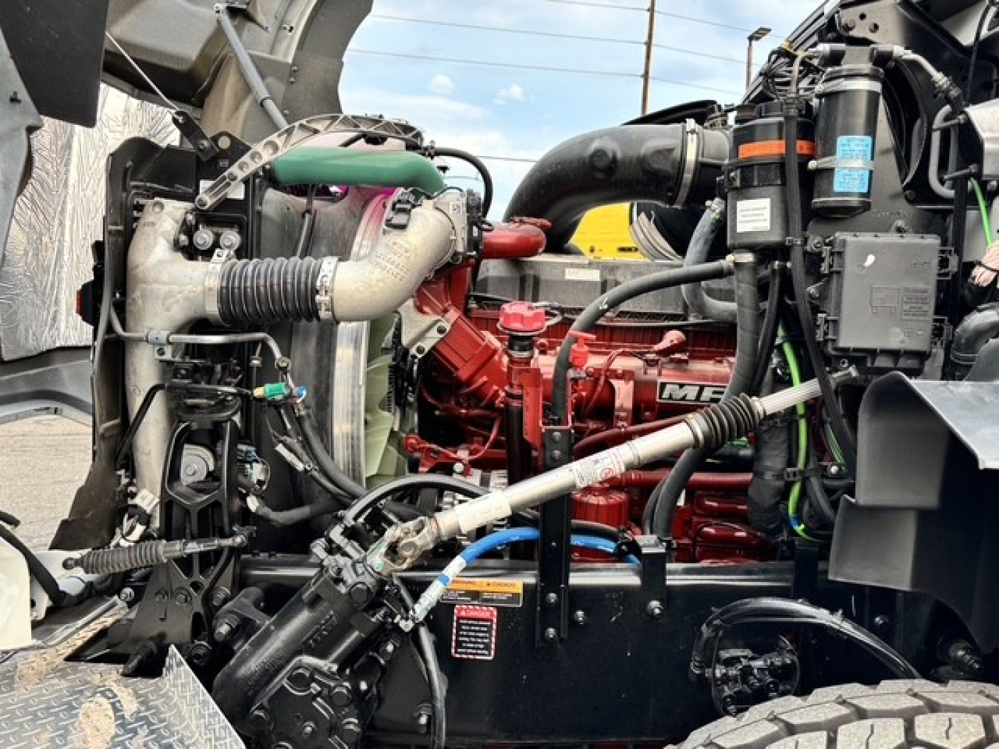 Mack GR64B engine bay showing red MaxxForce/Mack diesel engine with hoses, coolant reservoir, and surrounding components.