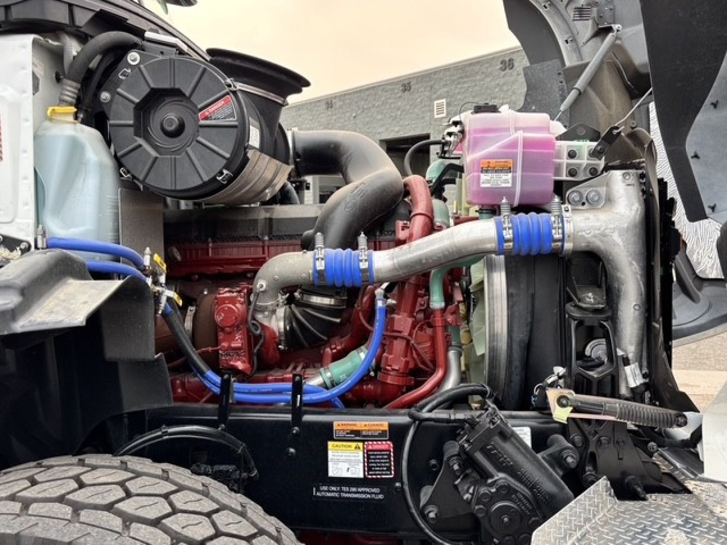 Mack GR64B engine bay showing red engine block, blue hoses, pink coolant reservoir, air intake assembly, radiator and belts.