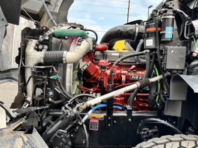 Mack GR64B engine bay showing red MaxxForce/Mack diesel engine with hoses, coolant reservoir, and surrounding components.