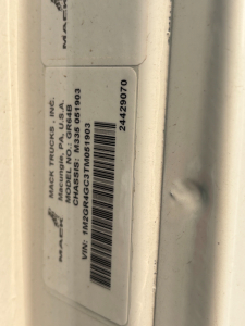 Label on Mack truck door frame showing MACK TRUCKS INC, MODEL GR64S? or GR64? with VIN 1M2GR4GC3TM051903 and stock 691?003; barcode; other text rotated.