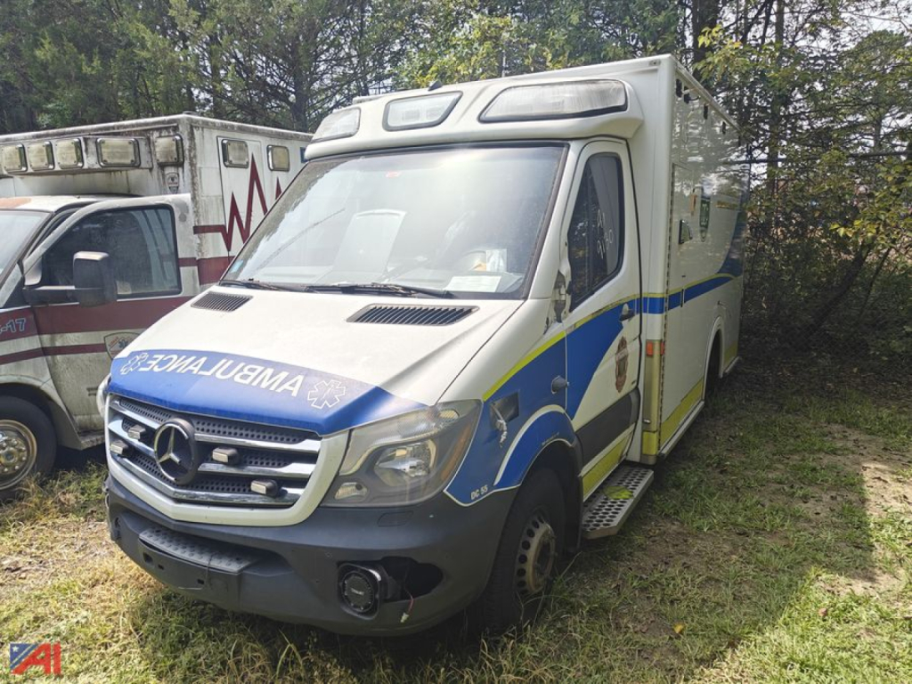 Ambulance vehicle from front-right showing Mercedes-Benz Sprinter with blue and yellow graphics, emergency lights, and 'AMBULANCE' reversed on front grille area.