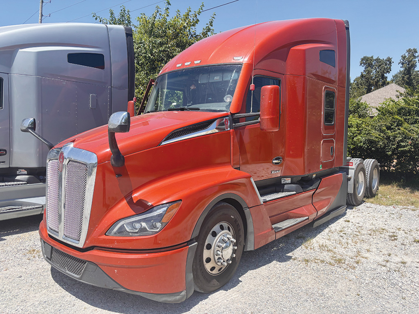Kenworth T680 Next Gen tractor in red, front-left view, with sleeper cab and chrome grille, parked on gravel.