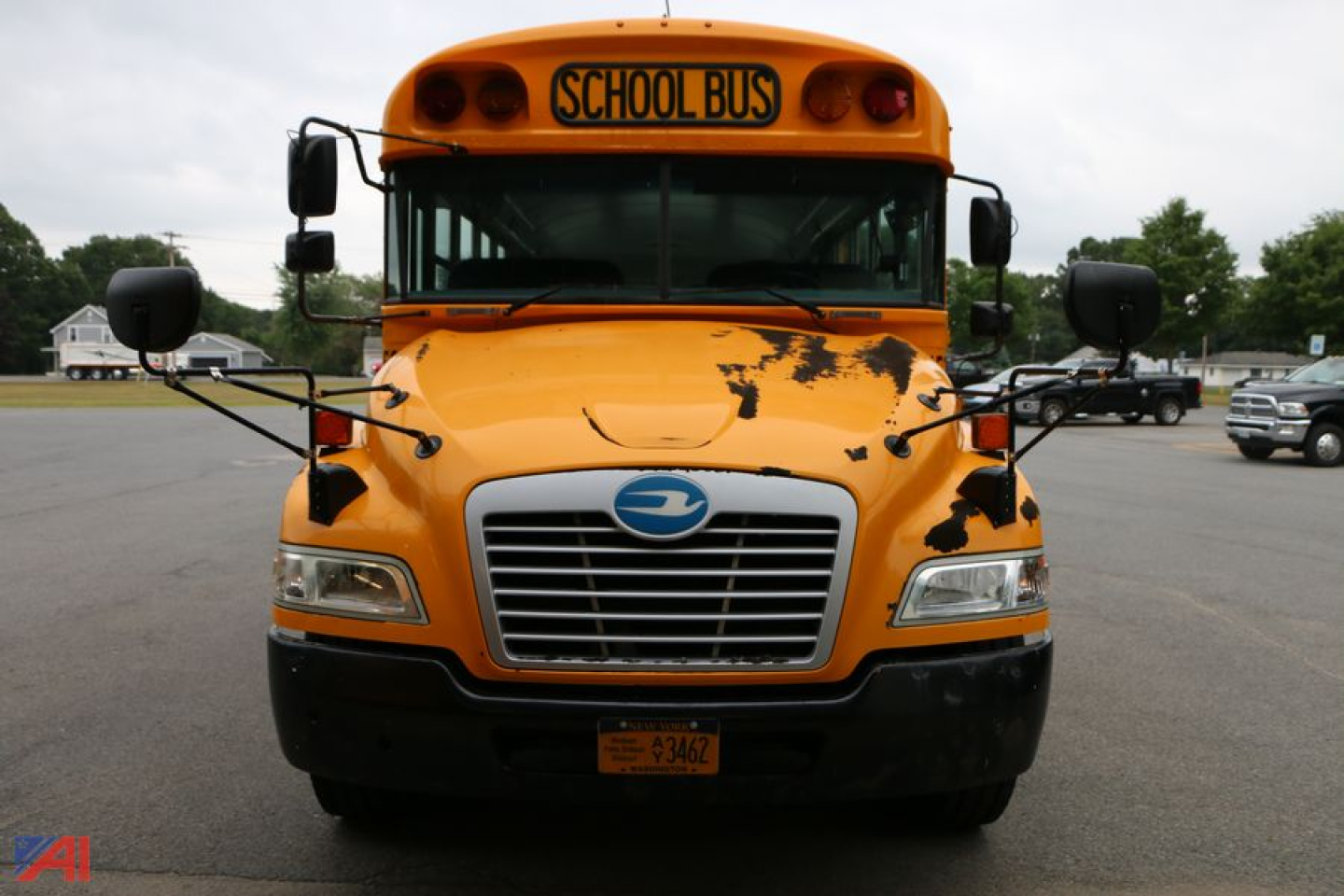Front view of a yellow/blue school bus with a Schhol Bus sign on top, front grille, headlights, and side mirrors.
