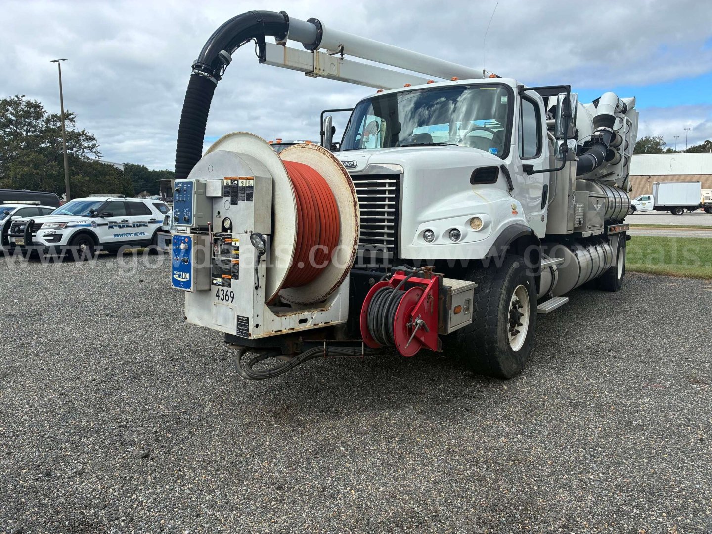White Freightliner M2 106 vacuum truck with large hose reel at front and long boom; visible labels: blue information plate, red hose reels, black intake hose, multiple tanks and equipment along sides.