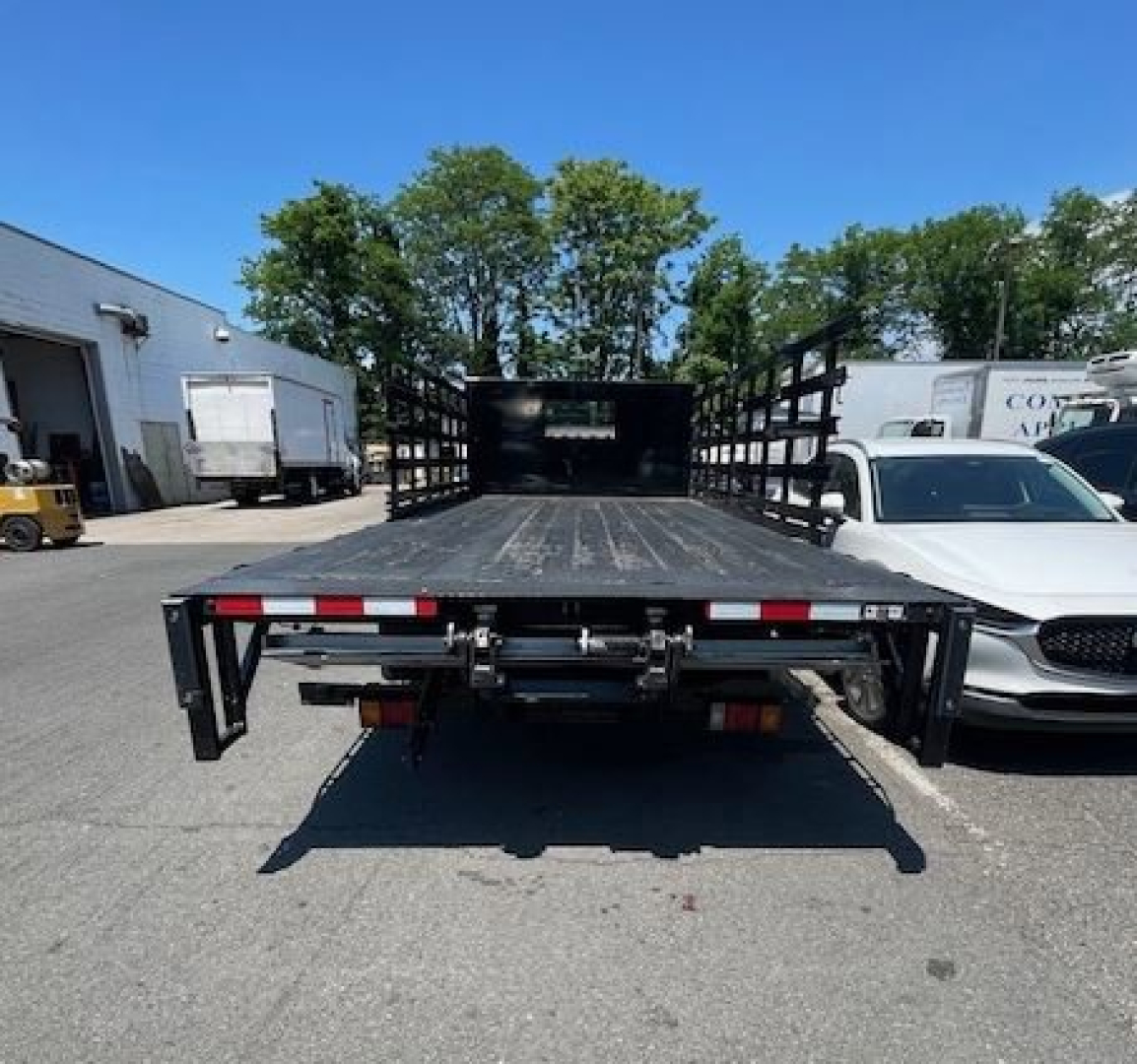 Isuzu NPR HD flatbed truck rear view showing open cargo bed with ramp and linkage, surrounding vehicles and lot.