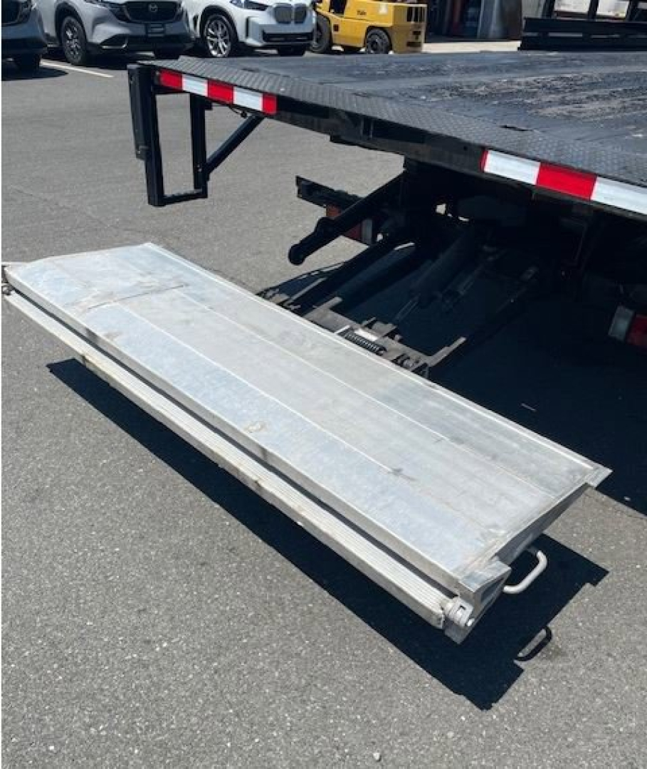 Isuzu NPR HD truck bed with a deployed aluminum folding ramp, rear view showing truck chassis and loading ramp mechanism.