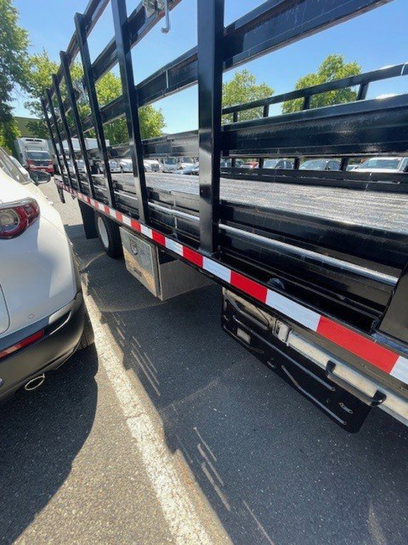 Isuzu NPR HD truck side showing metal stake bed with black bars and reflective red-white safety tape along the side.