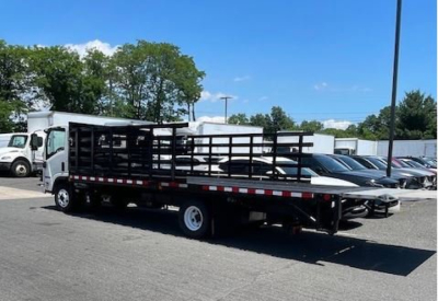 Isuzu NPR HD box truck with flatbed stake bed, side rails, and cab in foreground; multiple parked vehicles in background; clear sky.