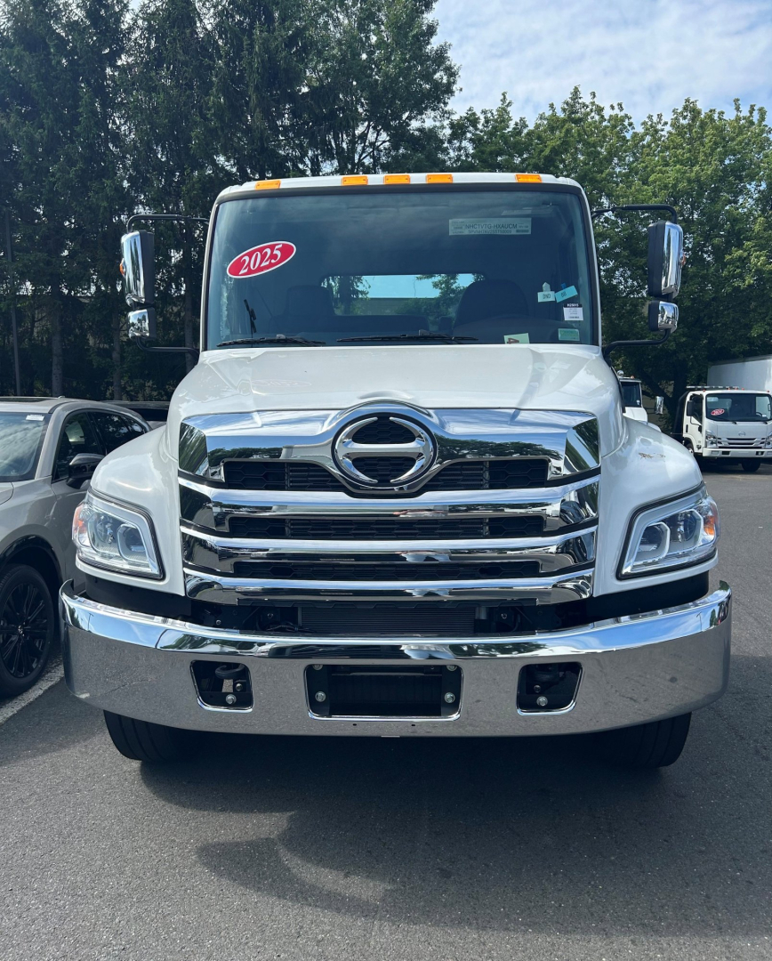 Front view of a white 2025 Hino L6 tow truck showing chrome grille, bumper, and Hino emblem, with windshield sticker and side mirrors.