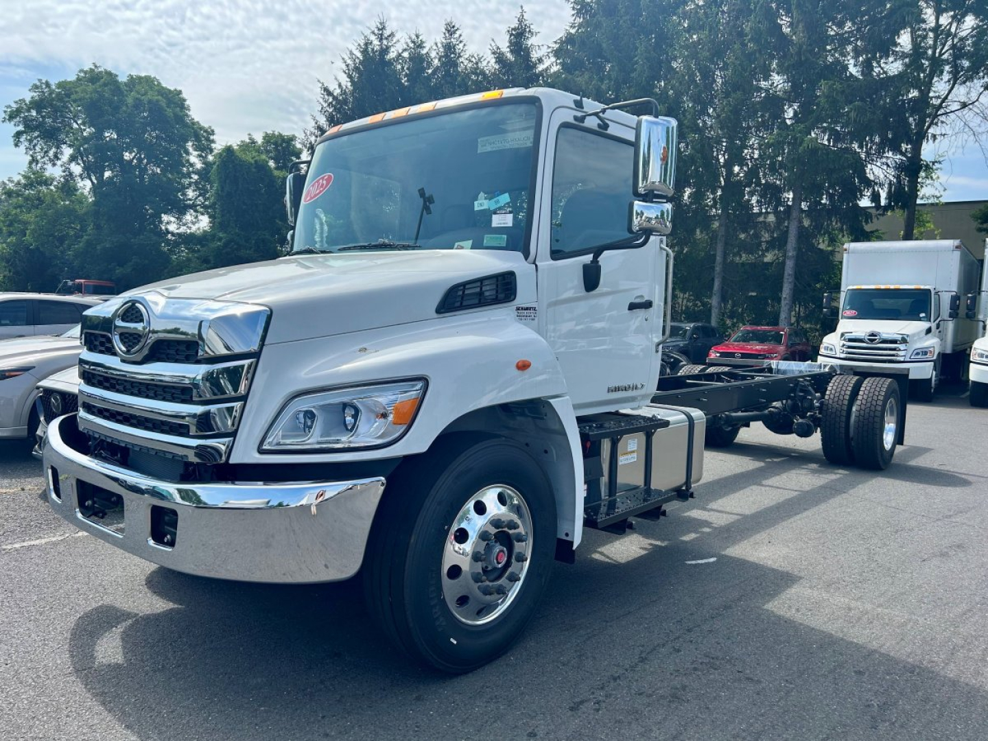 Truck cab and chassis of a 2025 Hino L6 tow truck, white exterior, chrome grille, dual rear axles, wheel rims visible, side mirrors, front bumper; shows cab and empty tow-bed chassis.