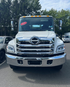 Front view of a white 2025 Hino L6 tow truck showing chrome grille, bumper, and Hino emblem, with windshield sticker and side mirrors.
