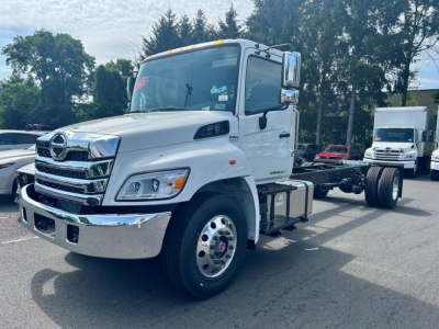Truck cab and chassis of a 2025 Hino L6 tow truck, white exterior, chrome grille, dual rear axles, wheel rims visible, side mirrors, front bumper; shows cab and empty tow-bed chassis.