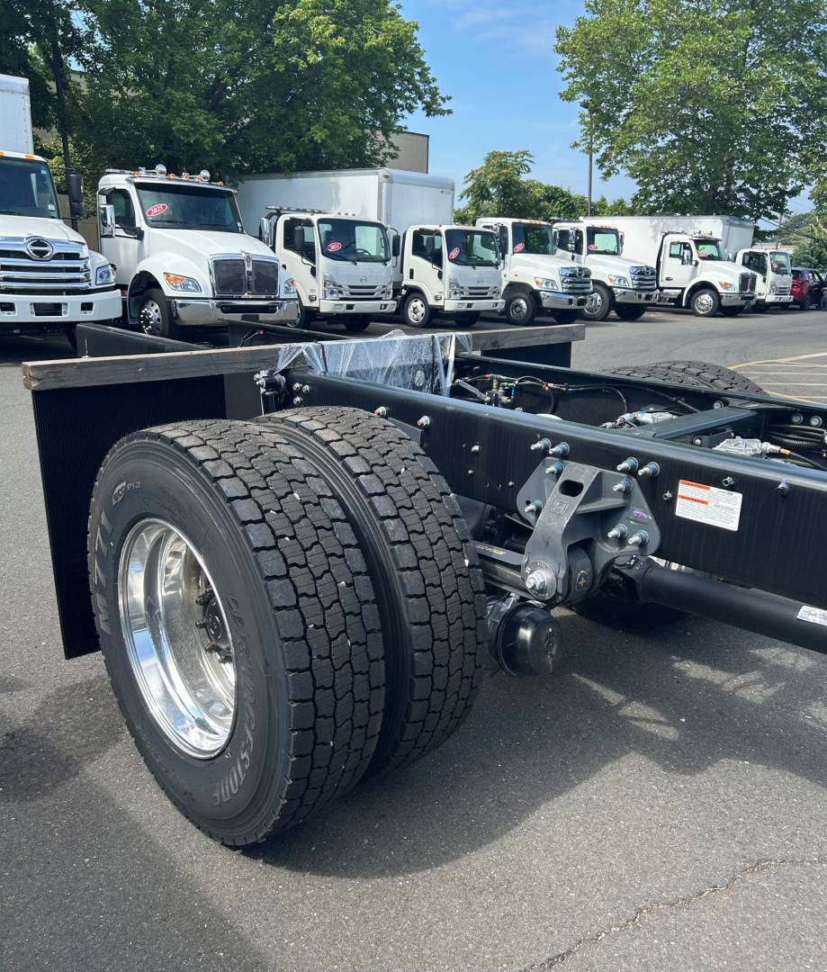 Truck chassis rear wheel and frame showing dual rear tires and chassis components with a line of cab chassis trucks in the background.