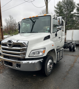 Hino L6 cab chassis truck front view showing white cab and chrome grille with Hino emblem, headlights, side mirrors, and front bumper.