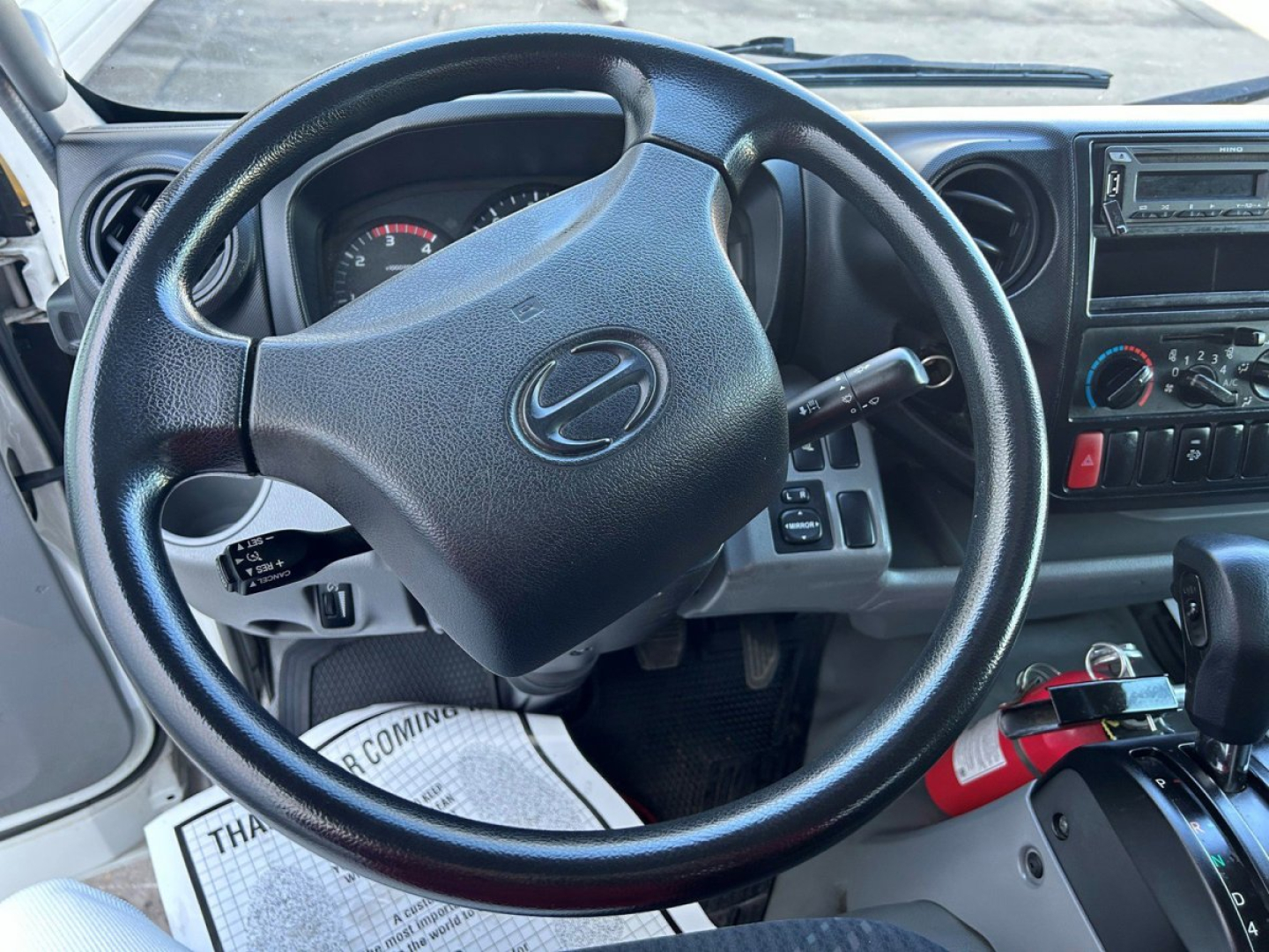 Steering wheel and dashboard of a 2018 Hino 155 box van truck interior showing steering wheel, gauges, center console, and gear lever.