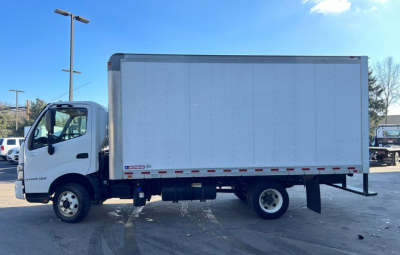 Box truck side view showing a white Hino box body with cab in front and gray cargo area; chassis and wheel wells visible.
