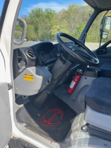 Truck cab interior of a 2018 Hino 155 showing steering wheel, dashboard, driver seat, and fire extinguisher mounted near door; floor mat with red logo.