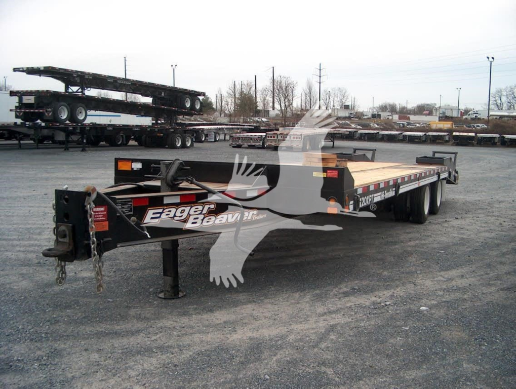 Trailer showing Eager Beaver logo on front, flatbed with wood deck and dual rear axles in a vehicle yard.