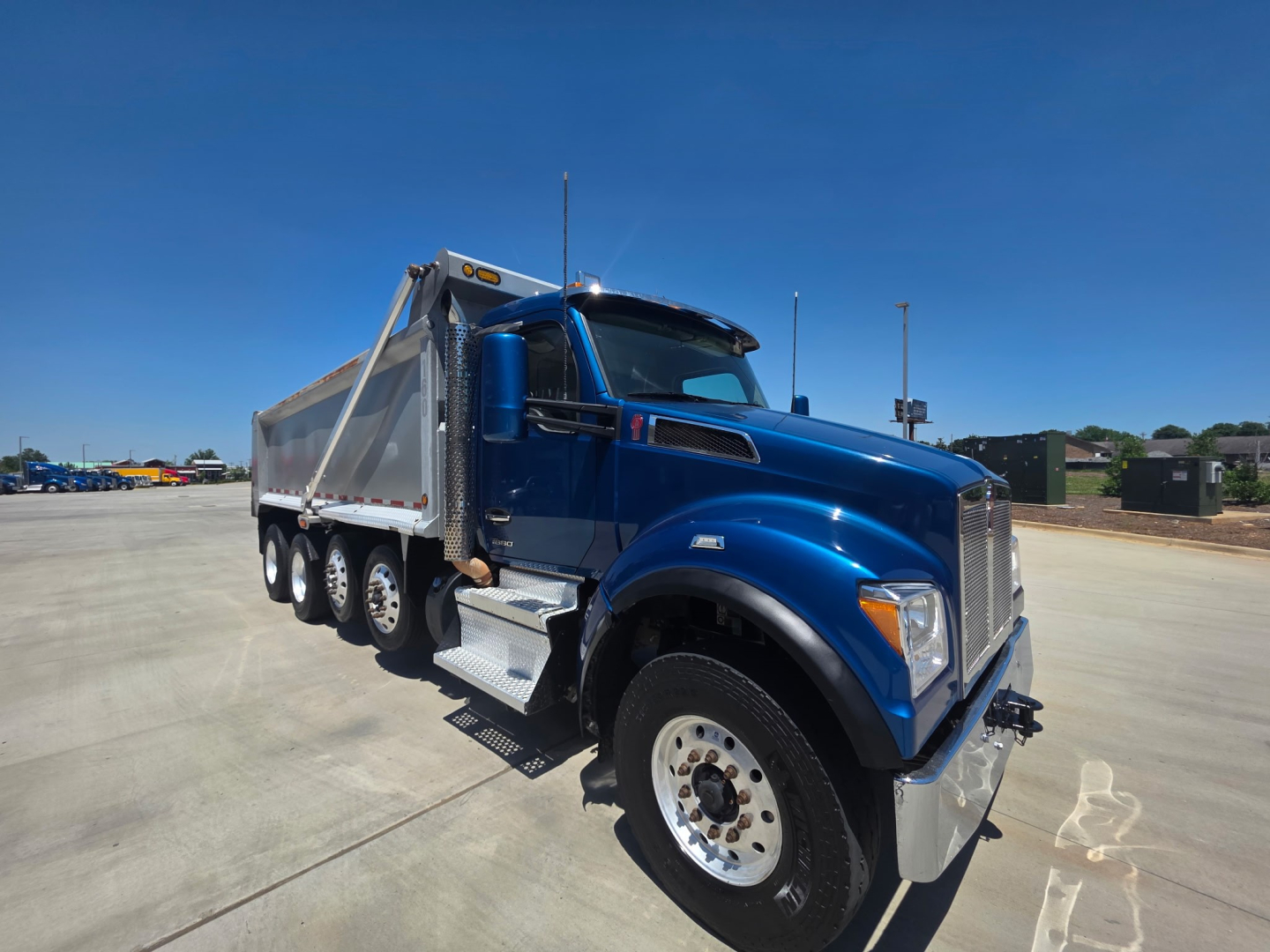 Blue 2023 Kenworth T880S dump truck shown at an angle with dump bed, front grille, large side mirrors, and multiple rear axles.