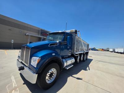 Kenworth T880S dump truck in blue exterior, front-left view showing large radiator grille, dual tandem rear axles, chrome bumper, and dump body. No visible text readable.