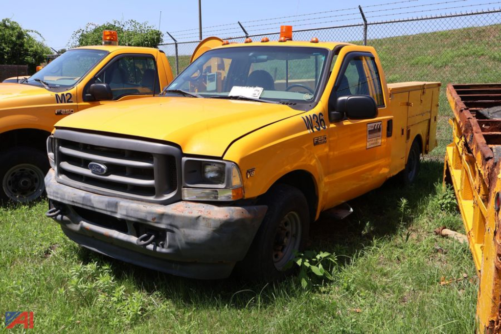 Ford F-250 Super Duty utility truck in yellow with utility bed, front bumper dented, parked on grass next to another yellow service truck.