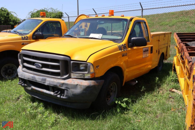 Ford F-250 Super Duty utility truck in yellow with utility bed, front bumper dented, parked on grass next to another yellow service truck.
