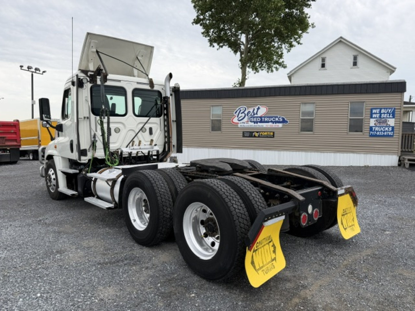 Freightliner Cascadia CA125DC truck tractor rear chassis and dual rear axles with mud flaps, yellow caution signs, and a beige service building in background.