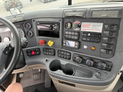 Cab interior of a Freightliner Cascadia showing dashboard controls, gauges, steering wheel, and center console with various switches.