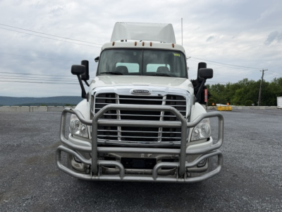 Front view of a white 2018 Freightliner Cascadia CA125DC semi truck with a heavy grille guard and bumper in a gravel lot.