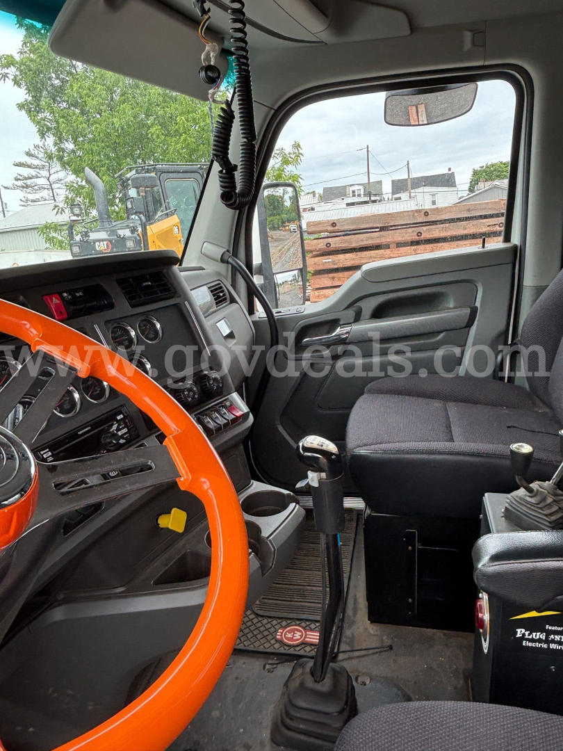 Kenworth T880 truck interior showing steering wheel, dashboard, gear shifter, and passenger seat; windshield with rearview mirror; hanging lanyard and cords; floor mat and control panels; label text not clearly legible.